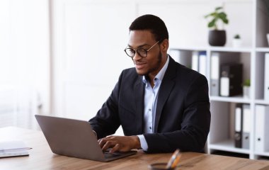 Resmi takım elbiseli neşeli genç girişimci dizüstü bilgisayarla çalışıyor, gözlük takıyor, ofis içi, fotokopi odası. Başarılı bin yıllık Afro-Amerikan iş adamı iş projesi üzerinde çalışıyor.