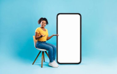 Mobil uygulama ya da web sitesi tasarımı için tasarlanmış, mavi stüdyo arka planı olan boş akıllı telefonun yanında oturan Afrikalı güzel bir bayan. Çevrimiçi reklamınız için boşluk