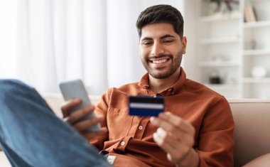 İnternet Mobil Alışveriş Konsepti. Cep telefonu tutan, kredi kartı kullanan, oturma odasındaki kanepede oturmuş, internetten bir şeyler alan, web mağazasından hediyeler alan bir Orta Doğulu.