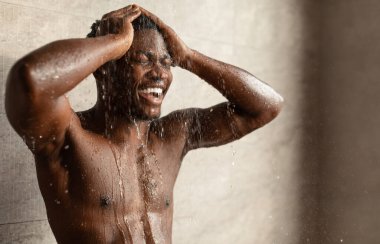 Duş alan Afrikalı Amerikalı adam, yıkanılan suyun altında dikiliyor ve kafasını modern banyoda yıkıyor. Erkekler için saç bakımı ve vücut bakımı kavramı. Seçici Odaklanma