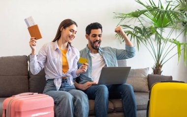 Excited tourists couple booking hotel room online, using laptop and credit card, sitting on sofa with suitcases, passports and tickets, ready for vacation trip