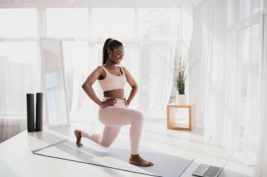 Genç siyah bir dişi, parlak ve boş bir odada hamlelerini yaparken spor rutinine odaklanır. Ev ödevi sırasında gücünü arttırmak için çevrimiçi bir dersi takip ediyor..