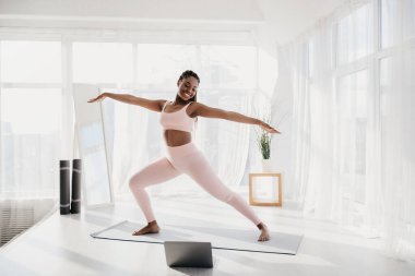 Neşeli genç siyah kadın savaşçı pozuyla evde yoga yapmaktan hoşlanıyor. Işıl ışıl iç mekanda geniş pencereler ve sakin bir atmosfer var. Egzersiz alışkanlığını geliştiriyor..