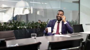 Pozitif Afro-Amerikan genç iş adamı, pahalı mavi takım elbiseli, ofisinde çalışıyor, kişisel asistanı veya iş ortağıyla telefon görüşmesi yapıyor ve gülümsüyor,