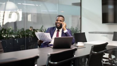 Gülümseyen Afro-Amerikalı takım elbiseli bir işadamı asistanı ya da iş ortağıyla telefon görüşmesi yapıyor, dizüstü bilgisayarının önünde oturuyor, elinde belgeler tutuyor, elinde fotokopi alanı olan bir panorama.
