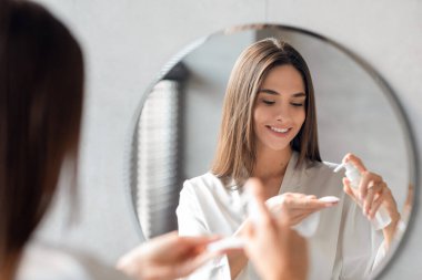 Genç bir kadın banyodaki aynanın yanında dikilirken pamuk pede temizlik jeli sürüyor. Yüzündeki makyajı kaldırıyor, yansımasına odaklanıyor..