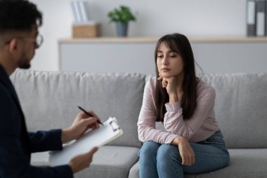 Orta doğulu bir kadın psikiyatrist ile terapi seansı yaparken üzgün görünüyor. Bir kanepede oturuyor, sakin ve profesyonel bir ortamda duygularını paylaşıyor..