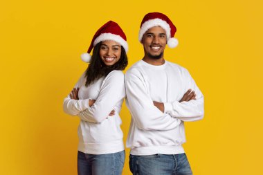 Sırt sırta verip gülümseyen ve Noel Baba şapkası takan iki genç yetişkin. Gündelik beyaz kazaklar giyiyorlar, neşeli bir tatil ruhunu canlı bir ortamda sergiliyorlar..