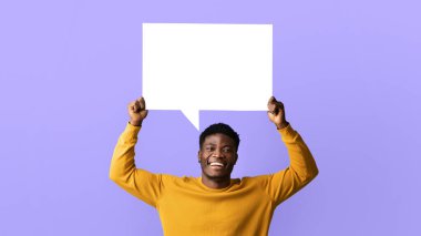 Genç bir adam kafasının üzerine boş bir işaret kaldırırken geniş bir şekilde gülümser. Canlı, mor bir arka planın önünde duruyor, parlak ve neşeli bir atmosfer sergiliyor..