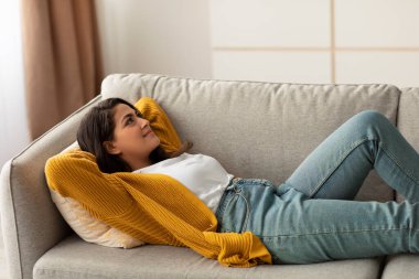 Sakin bir Arap kadın, elleri başının arkasında rahat bir kanepede uzanıyor, dinlendirici bir anın tadını çıkarıyor. Oturma odası aydınlık ve davetkar. Rahatlamak ve hayal kurmak için ideal bir yer..