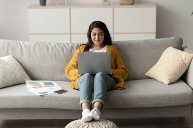 Genç bir Arap kadın serbest yazar bilgisayarını çalışmak ya da evde öğrenmek için kullanıyor. Kanepede rahatça oturuyor ve daktilo yazarken odaklanmış ve mutlu görünüyor..