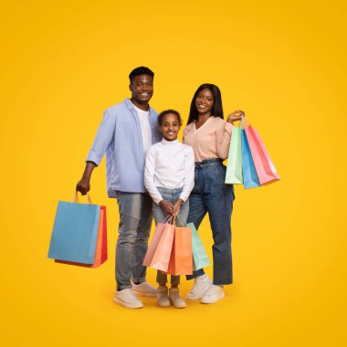 Neşeli bir aile, canlı alışveriş torbaları taşıyan bir arada durur. Parlak sarı bir arka planda mutlu bir şekilde gülümserler. Alışverişin ve birlikte zaman geçirmenin keyfini yakalarlar..
