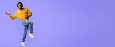 Sarı kazaklı ve mavi kotlu bir adam canlı mor bir arka planda neşeyle dans ediyor. Kulaklık takıyor ve müzikten hoşlanıyor gibi görünüyor. Mutluluğunu gösteriyor..