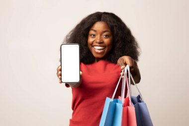 Mutlu bir Afrikalı Amerikalı kadın elinde canlı alışveriş torbaları tutarken beyaz ekranlı bir akıllı telefon sergiliyor..