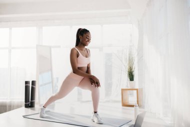 Milenyum siyahi kadın, online bir çalışmanın ardından parlak bir odada hamle yapıyor. Bilgisayarı kullanarak spor yapmaya odaklanıyor. Kilitleme sırasında evde egzersiz yapıyor..