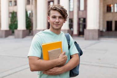 Üniversite binasının önünde elinde not defterleri ve sırt çantası olan genç bir erkek öğrenci duruyor. Açık renk bir gömlek giyiyor ve derse hazır görünüyor..