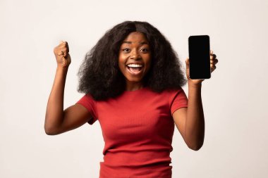 Neşeli bir Afrikalı Amerikalı kadın beyaz bir arka plana karşı duruyor, heyecanlı bir şekilde yumruğunu kaldırıyor ve boş bir akıllı telefon tutuyor. Şu anda mutluluğu ve zaferi ifade ediyor..