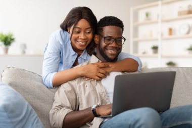 Genç siyahi çift, eşi dizüstü bilgisayarı üzerinde çalışırken onu arkadan kucaklarken gülümsüyor. Sıcak bir ev ortamında rahat bir kanepede oturuyorlar..