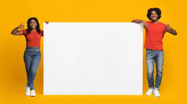 İki birey parlak sarı bir zemine karşı duruyor, her biri büyük boş bir işaret taşıyor. Gülümsüyorlar ve başparmak kaldırıyorlar, neşeli ve pozitif bir atmosfer yayıyorlar..
