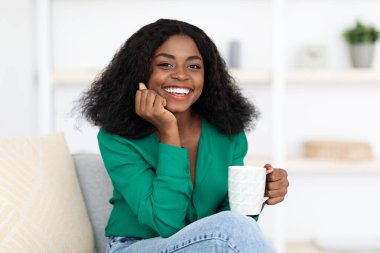 Kıvırcık saçlı neşeli bir kadın elinde bir kupa tutuyor ve şık bir oturma odasında rahatça otururken gülümsüyor. Doğal ışık onun etrafındaki boşluğu aydınlatır..