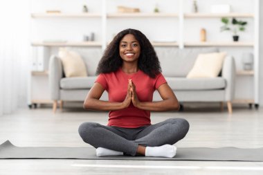 Neşeli bir kadın oturma odasında yoga minderinin üzerinde elleri bağlı dua pozisyonunda oturur. Güneş ışığı boşluğu doldurur ve sıcak bir atmosfer yaratır..