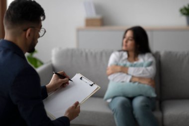 Üzgün genç bir Arap kadın kanepeye oturmuş, psikoloğuyla konuşurken yastığına sarılıyor. Psikiyatrist oturum sırasında not almaya odaklanmış..