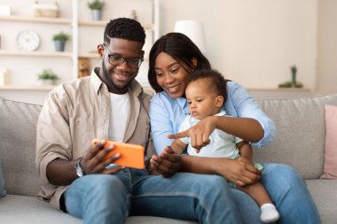 Genç Afrikalı Amerikalı ebeveynler evlerindeki bir kanepede oturur, küçük oğullarına cep telefonlarında komik bir eğitim uygulaması gösterirken gülümserler. Anne bebeği tutup, ekranı işaret ediyor..