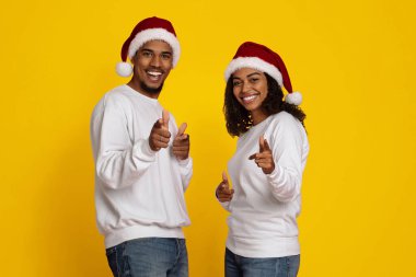 Neşeli bir çift Noel şapkaları ve günlük beyaz kazaklar giyerek kameraya poz veriyor. İkisi de tatil sezonunda mutluluk saçan parmaklarıyla işaret ediyor..