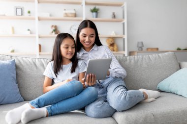 Genç bir Asyalı anne ve kızı bir kanepede rahatça oturuyorlar, video izlemek ve İnternet 'i keşfetmek için dijital tablet kullanıyorlar. Cihazla uğraşırken gülümseme paylaşıyorlar..