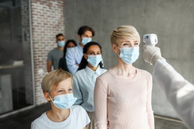 Farklı yaşlarda, koruyucu yüz maskeleri takan çok ırklı hastalar klinikte veya aşı merkezinde koronavirüse karşı aşılanmadan önce vücut ısısını elektronik termometreyle kontrol ediyorlar.