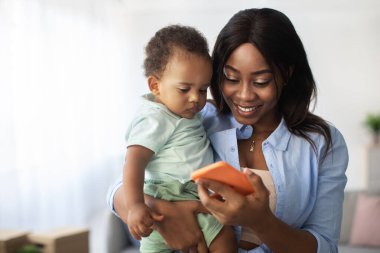 Neşeli bir Afrikalı Amerikalı anne, bebeğine evde akıllı bir telefon gösterirken onu kucağına alıyor. Eğitimsel bir uygulamanın tadını çıkarıyorlar, bağlanıyorlar ve birlikte eğleniyorlar..