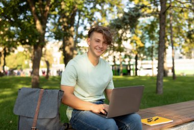 A young man sits on a park bench with a laptop, focused on his work. He smiles at the camera while enjoying the warm sunlight and natural surroundings.