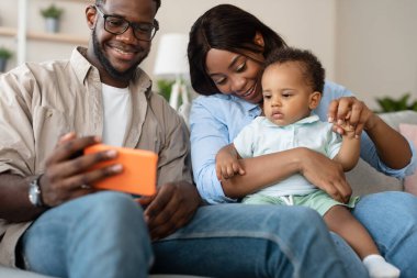 Neşeli bir Afrikalı Amerikalı aile koltukta birlikte oturur, cep telefonu kullanırken bir anı paylaşırlar. Bir video görüşmesi yapmak ve fotoğraflara bakmak gibi aktivitelerle meşguller..
