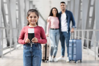 Havaalanı terminalinde seyahat biletlerini tutan genç bir kız gülümsüyor. Ailesi bagajlarıyla arka planda duruyor, aile maceralarına hazırlanıyorlar..