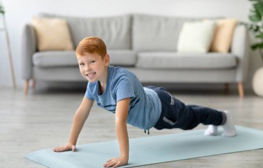 Oturma odasında kızıl saçlı bir çocuk yoga minderinde şınav çekiyor. Aktif kalmayı ve rahat bir atmosferle çevrili sağlıklı bir yaşam tarzını desteklemeyi seviyor..