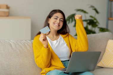 Heyecanlı genç Arap kadın koltuğunda oturuyor bir elinde kredi kartı ve önünde bir dizüstü bilgisayarla. Çevrimiçi piyangodan büyük bir ikramiye kazandı ve evden alışveriş çılgınlığının tadını çıkarıyor..