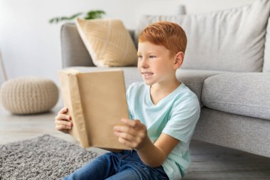 Kızıl çocuk evde yerde oturmuş, mutlu bir şekilde kitap okuyor. Heyecan verici hikayeler keşfederken gülümsüyor. Konforlu ortam, sayfalardaki maceradan zevk almasını sağlıyor..