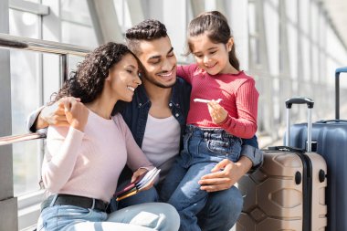 Üç kişilik bir aile havaalanında neşeli bir anı paylaşıyor, gülümseme ve kahkahalarla seyahat kartlarını tutuyorlar ve bavullarını kontrol ediyorlar. Yolculukları için heyecanlı görünüyorlar..