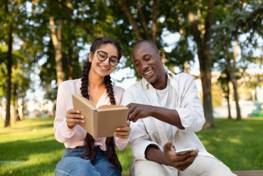 İki arkadaş yeşil bir parkta bir bankta oturuyor, birlikte gülüyor ve gülüyorlar. Biri kitap okurken diğeri sayfadaki bir şeye işaret ediyor..