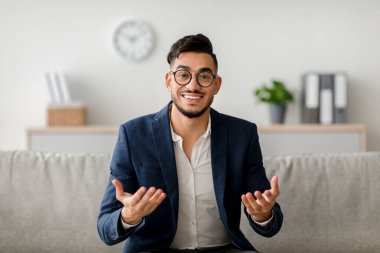 Genç bir ortadoğu psikoloğu rahat bir koltukta oturur, gülümser ve el kol hareketi yapar. Modern bir ofis ortamında psikoterapi seansına katılıyor..