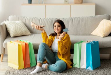 Orta Doğulu bir kadın, elinde bir kredi kartı ve yeni bir ayakkabıyla, elinde renkli alışveriş torbalarıyla yerde oturuyor ve rahat evinde online alışverişin keyfini çıkarıyor..