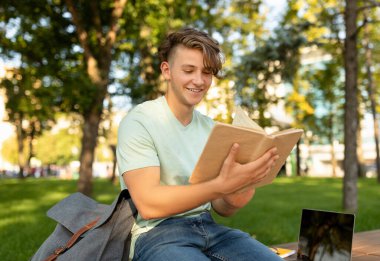 Genç bir adam parkta bir bankta oturuyor, etrafı ağaçlarla çevrili, elinde kitabı okurken mutlu bir şekilde tutuyor. Yakınlarda bir dizüstü bilgisayar duruyor, rahatlatıcı atmosfere ekleniyor..