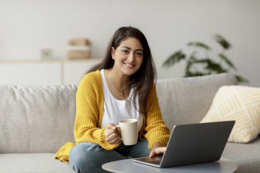 Neşeli Arap kadın serbest yazar evdeki kanepede oturuyor. Dizüstü bilgisayarını kullanırken elinde bir fincan kahve tutuyor, rahat ve davetkar bir yerde işinin tadını çıkarıyor..