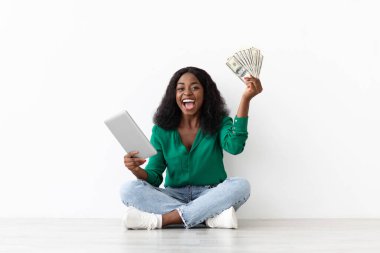 Neşeli genç bir kadın yeşil bir bluzla yerde oturuyor, bir elinde para, diğer elinde tablet var. O, başarısının heyecanını parlak bir ortamda ifade eder..