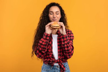Neşeli bir bayan lezzetli bir burgeri seve seve tutuyor ve kokluyor. Ekose bir gömlek giyer ve canlı sarı bir arka plana yaslanır. Atıştırmalıklarının tadını çıkarmaya hazırdır..