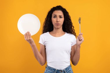 Kıvırcık saçlı genç bir kadın bir çatal ve boş bir tabak gösteriyor, parlak sarı stüdyo arka planında üzüntüsünü ifade ediyor. Diyet ve açlığı yüzünden hayal kırıklığına uğramış..