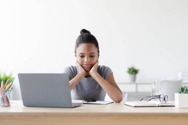 Yorgun genç siyah bir kız, dizüstü bilgisayarına bakarken çenesini ellerinin üzerinde dinlendiriyor. Düşünceli görünüyor, rahat bir evde online çalışmalarına ara veriyor..