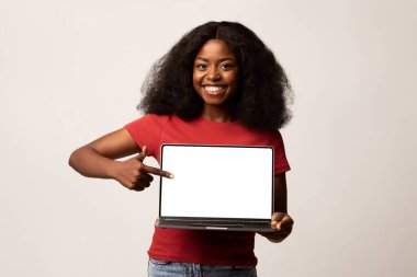 Neşeli siyah bir kadın boş bir dizüstü bilgisayarı işaret ederken gülümsüyor. Çevrimiçi içerik veya reklam olanaklarını destekleyen hafif bir arkaplan karşısında duruyor..