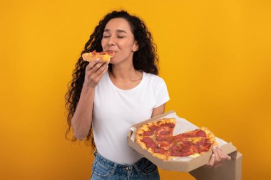 Heyecanlı genç bayan pizza kutusunu tutarken bir dilim pizzayı ısırıyor. Neşeli bir stüdyoda canlı sarı bir geçmişi var, lezzetli bir ziyafetin tadını çıkarıyor..