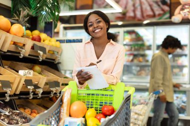 Genç bir kadın alışveriş arabasının yanında taze meyve ve sebzelerle dolu olarak dururken bir panoya alışveriş listesi yazarken gülümsüyor..